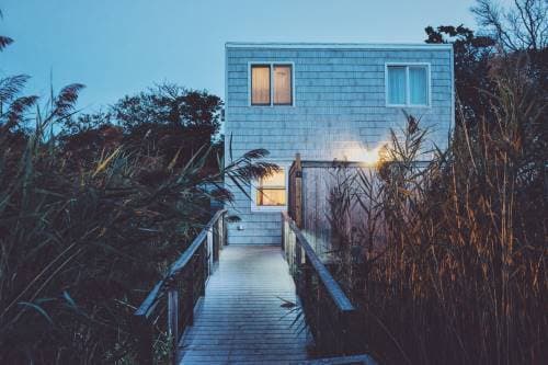 A house with light gray siding, with two rectangular windows on the upper floor and one on the ground floor. The house is located on a lot with tall vegetation, and a wooden deck with handrails leads to the front door. The scene is illuminated by soft light, suggesting late afternoon or early evening.