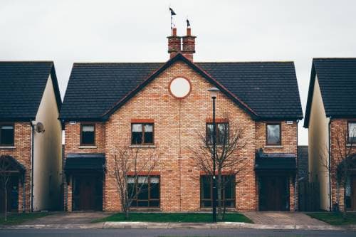 A row of brick houses with black roofs, two chimneys and rectangular windows. The houses have a symmetrical design and are separated by a small garden. The street in front of the houses is paved and there is a lamppost next to it.