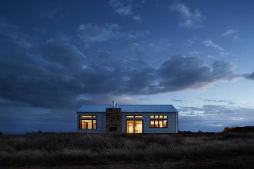 Front view of a single-family home with a modern design and straight lines. The house is situated on a flat lot with tall grass and a night sky with thick clouds. Lights on in the windows create a cozy atmosphere.