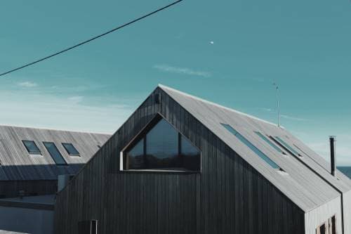 A contemporary house with a dark wood-clad facade and a pitched roof. The roof features several glass windows, providing natural light to the interior. In the background, a clear blue sky with a few clouds serves as a backdrop for the building.