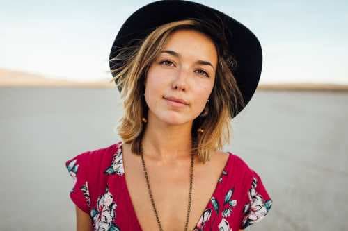 Blonde woman wearing a hat, necklace and floral blouse, there seems to be a sea behind her